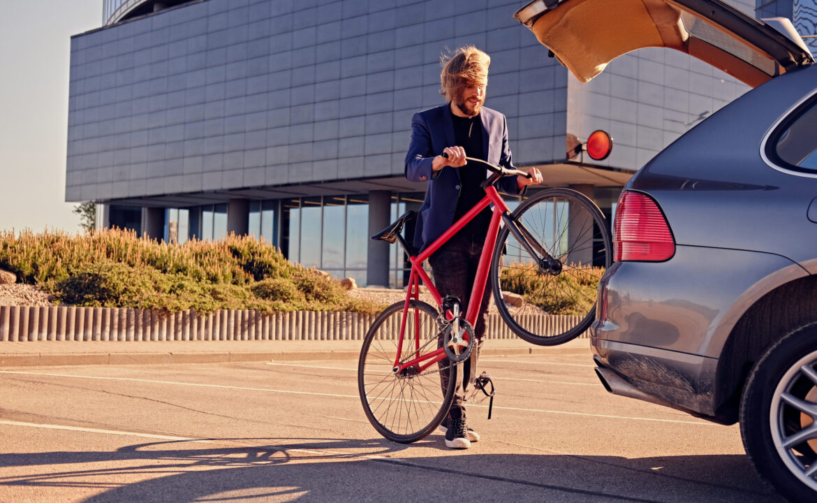 A man holds fixed bicycle near the car with open trunk.
